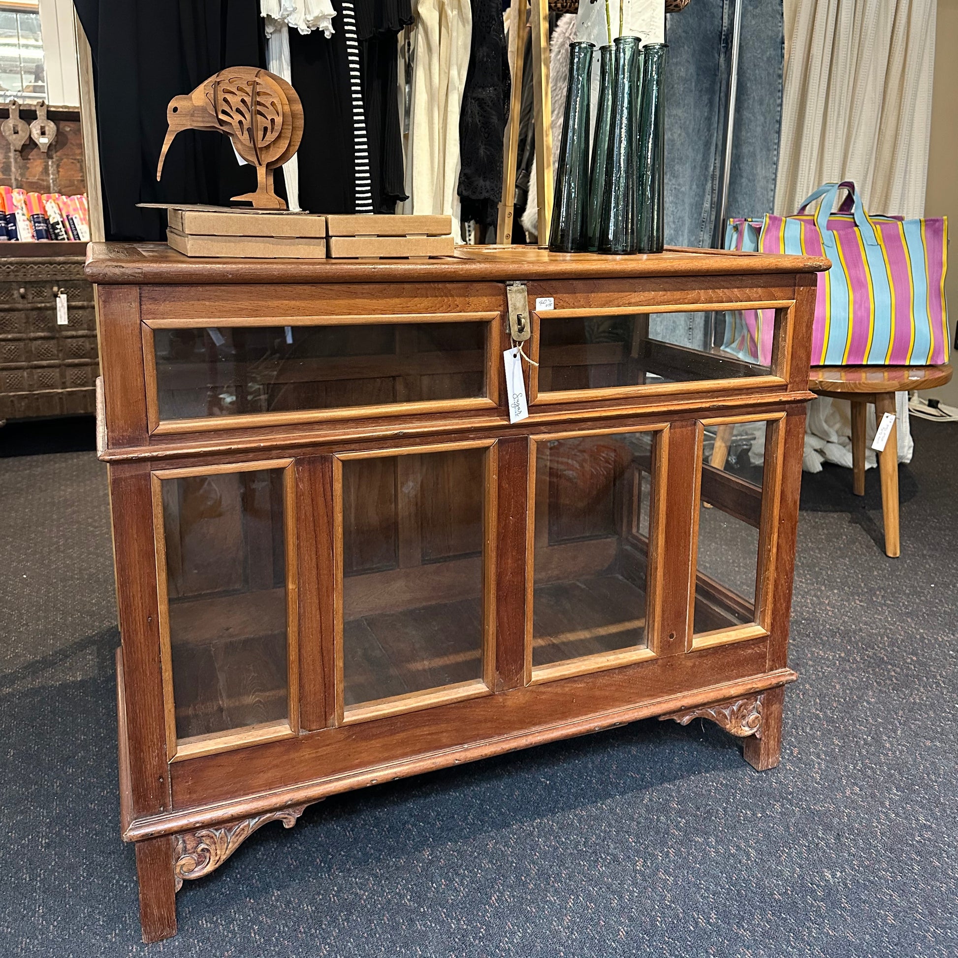 A vintage wooden display cabinet with glass doors and ornate carved legs, photographed in a retail store setting.